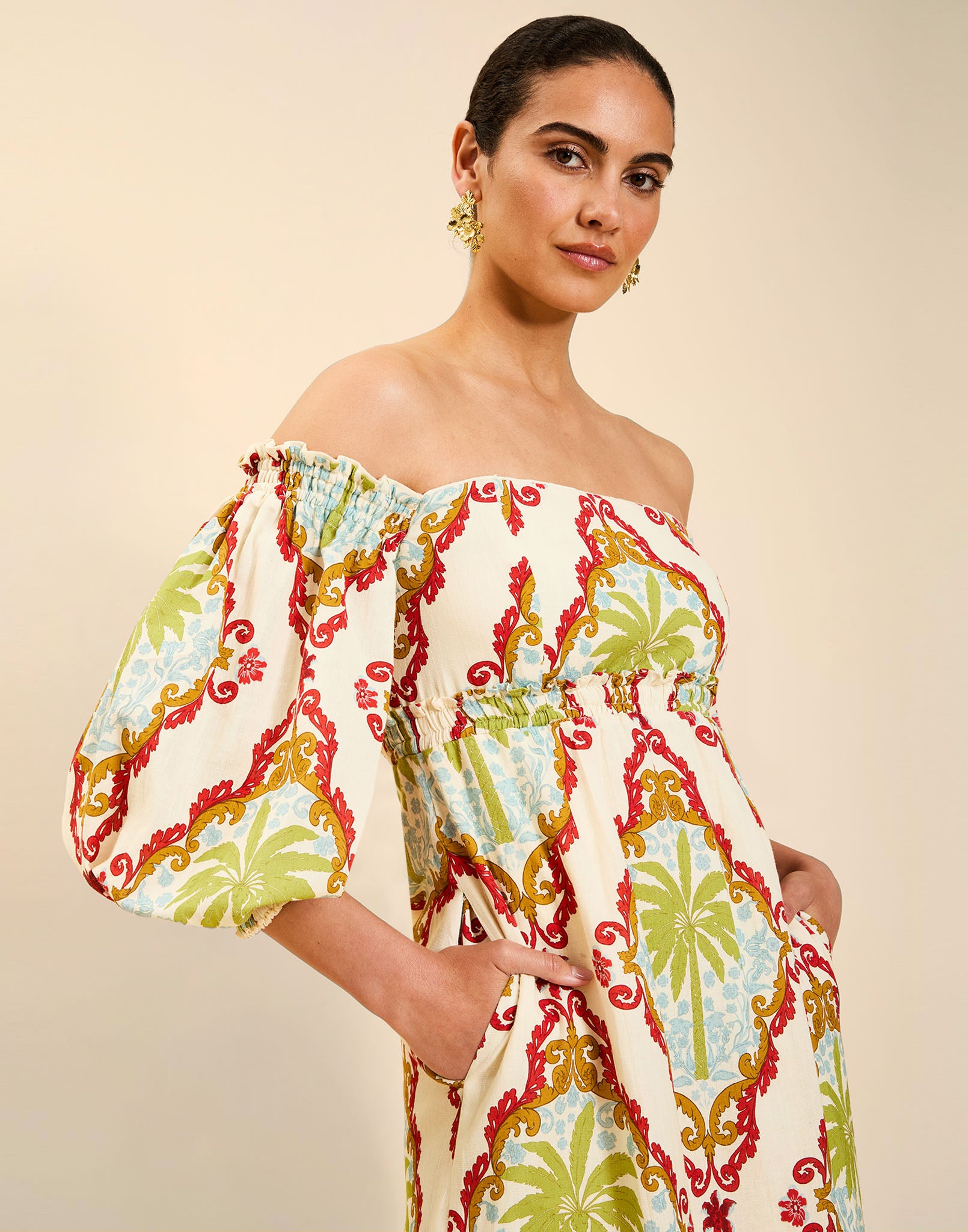 Woman wearing the Mestiza Benny Midi Dress in Mediterranean Sage print, featuring an off-the-shoulder neckline, empire waist, balloon sleeves, A-line skirt, and breathable linen fabrication.