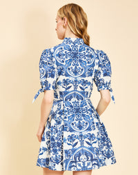 Woman wearing the Mestiza Brooklyn Mini Dress in Azulejo Navy print, featuring a mandarin collar, fitted waist, tulip skirt, elbow-length puff sleeves, tie cuffs, and cotton poplin fabrication.