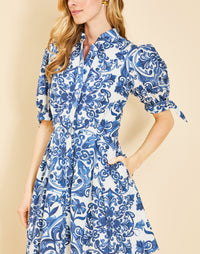 Woman wearing the Mestiza Brooklyn Mini Dress in Azulejo Navy print, featuring a mandarin collar, fitted waist, tulip skirt, elbow-length puff sleeves, tie cuffs, and cotton poplin fabrication.