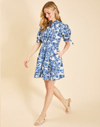 Woman wearing the Mestiza Brooklyn Mini Dress in Azulejo Navy print, featuring a mandarin collar, fitted waist, tulip skirt, elbow-length puff sleeves, tie cuffs, and cotton poplin fabrication.