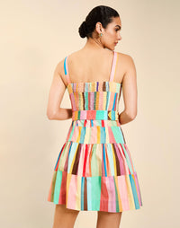 Woman wearing the Mestiza Castellano Mini Dress in Southwest Sunset print, featuring a drop-waist silhouette, fitted bodice with princess seams, adjustable straps, gathered skirt, coordinating belt, and cotton poplin fabrication.