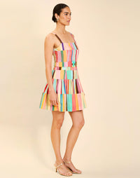 Woman wearing the Mestiza Castellano Mini Dress in Southwest Sunset print, featuring a drop-waist silhouette, fitted bodice with princess seams, adjustable straps, gathered skirt, coordinating belt, and cotton poplin fabrication.