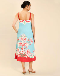 Woman wearing the Mestiza Galena Tea Length Dress in La Botanica Blue print, featuring a column silhouette, straight-across neckline, adjustable straps, side seam pockets, and cotton poplin fabrication.