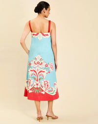 Woman wearing the Mestiza Galena Tea Length Dress in La Botanica Blue print, featuring a column silhouette, straight-across neckline, adjustable straps, side seam pockets, and cotton poplin fabrication.