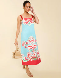 Woman wearing the Mestiza Galena Tea Length Dress in La Botanica Blue print, featuring a column silhouette, straight-across neckline, adjustable straps, side seam pockets, and cotton poplin fabrication.