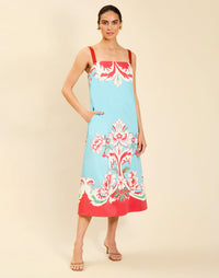 Woman wearing the Mestiza Galena Tea Length Dress in La Botanica Blue print, featuring a column silhouette, straight-across neckline, adjustable straps, side seam pockets, and cotton poplin fabrication.