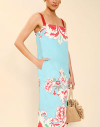 Woman wearing the Mestiza Galena Tea Length Dress in La Botanica Blue print, featuring a column silhouette, straight-across neckline, adjustable straps, side seam pockets, and cotton poplin fabrication.
