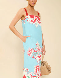 Woman wearing the Mestiza Galena Tea Length Dress in La Botanica Blue print, featuring a column silhouette, straight-across neckline, adjustable straps, side seam pockets, and cotton poplin fabrication.