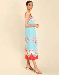 Woman wearing the Mestiza Galena Tea Length Dress in La Botanica Blue print, featuring a column silhouette, straight-across neckline, adjustable straps, side seam pockets, and cotton poplin fabrication.